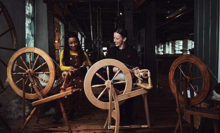 A lady sits with a craftsperson in front of 3 traditional wooden spinning wheels. The lady feeds the strands of wool to the craftsperson who is spinni